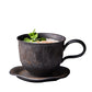 Black ceramic cup with handle on a matching saucer, containing a green leafy vegetable.