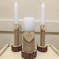 Three candles in rustic holders with wooden hearts and initials on a white surface.